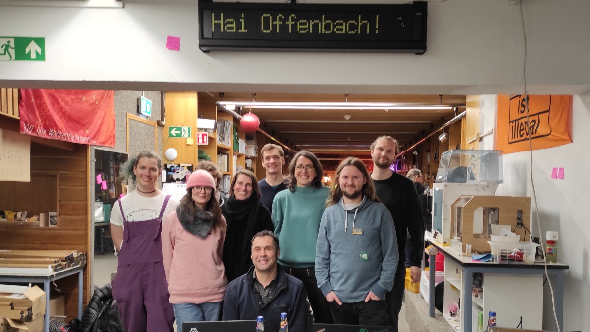Gruppenbild mit anwesenden Aktiven und der Besucher*innengruppe aus Offenbach unter dem Flipdot-Anzeiger