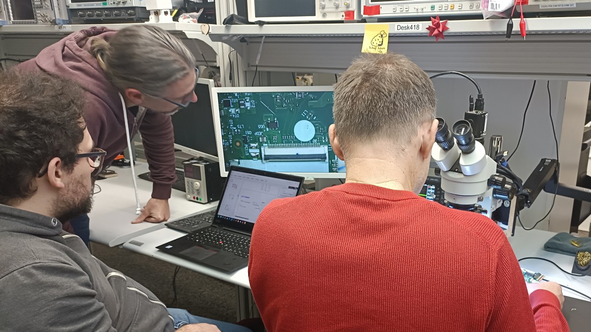 Im Elektroniklabor inspizieren drei Menschen eine Platine, die rechts unter dem Stereomikroskop liegt. In der Mitte ist das Videobild der Platine unter dem Mikroskop auf einem Bildschirm zu sehen.